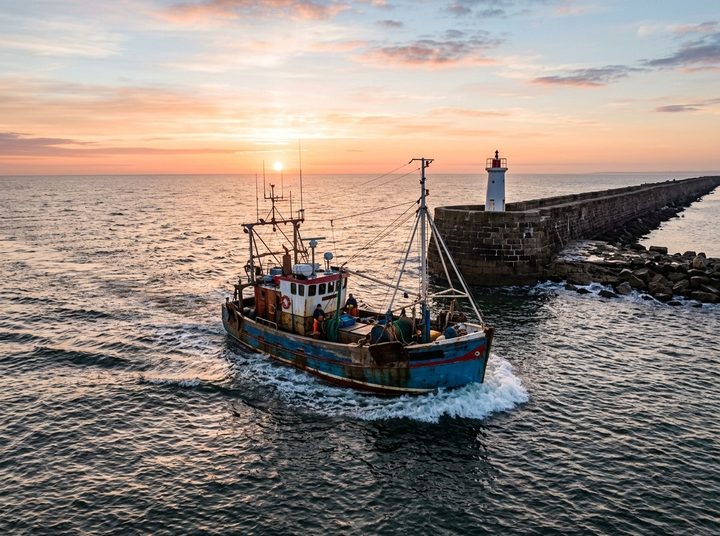 A fishing boat heading out past a harbour breakwater and lighthouse at sunrise